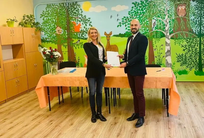 Zdjęcie o orientacji poziomej przedstawia dwie osoby stojące wewnątrz sali przedszkolnej, trzymające wspólnie białą kartkę dokumentu. Kobieta po lewej stronie ma na sobie czarną marynarkę, dżinsy i czarne botki, natomiast mężczyzna po prawej jest ubrany w ciemną marynarkę, bordowe spodnie i eleganckie buty. Stoją oni przed długim stołem przykrytym brzoskwiniowym obrusem, na którym widnieje bukiet kwiatów oraz przybory do pisania. Tło stanowi kolorowy mural przedstawiający leśne zwierzęta, drzewa oraz słońce, co nadaje wnętrzu edukacyjny charakter. Po lewej stronie kadru widoczna jest wysoka, drewniana szafka z roślinami doniczkowymi na blacie, a podłoga wykończona jest jasnymi panelami w kolorze drewna. Całość kompozycji jest symetryczna i dobrze oświetlona, idealnie nadając się do reportaży z wydarzeń oficjalnych w placówkach oświatowych.