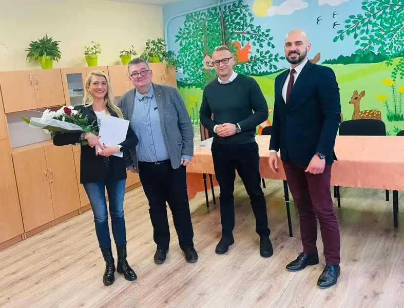 Zdjęcie wykonane w orientacji poziomej przedstawia grupę czterech osób stojących w jasnej sali przedszkolnej. Po lewej stronie stoi kobieta w czarnej marynarce i dżinsach, trzymająca bukiet kwiatów oraz białą teczkę, a obok niej znajdują się trzej mężczyźni w eleganckich strojach: od swetra z koszulą po garnitur z krawatem. W tle widać drewniane szafki, stół przykryty brzoskwiniowym obrusem oraz kolorowy mural na ścianie przedstawiający leśny krajobraz z sarną i drzewami. Podłoga w pomieszczeniu wyłożona jest jasnymi panelami drewnopodobnymi, a na górnych półkach szafek ustawiono kilka doniczek z zielonymi roślinami. Cała scena jest dobrze oświetlona, a postacie zajmują centralną część kadru, co sprawia, że zdjęcie ma charakter oficjalnego spotkania lub uroczystości w placówce edukacyjnej.