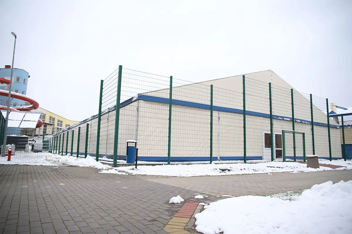Zdjęcie przedstawia dużą halę namiotową o jasnym kolorze, która jest otoczona wysokim ogrodzeniem z zielonej siatki panelowej. Budynek posiada dwuspadowy dach i wyraźne niebieskie akcenty konstrukcyjne, a przed wejściem widoczny jest pojedynczy niebieski kosz na śmieci. Ujęcie zostało wykonane z poziomu gruntu w formacie poziomym, ukazując plac wyłożony kostką brukową częściowo pokryty warstwą zalegającego śniegu. W lewej części kadru widać fragment kolorowej, czerwonej zjeżdżalni wodnej oraz nowoczesną bryłę sąsiedniego budynku, co sugeruje lokalizację przy obiekcie rekreacyjnym lub aquaparku. Całość utrzymana jest w chłodnej, zimowej tonacji z niebem zasnutym grubą warstwą jasnych chmur.